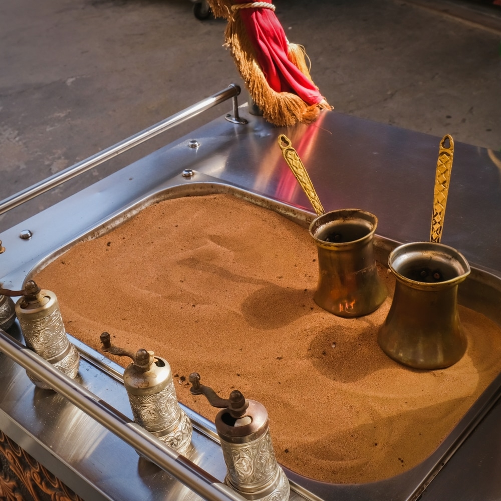 Two Cezve and hand mills with authentic Turkish coffee on hot sand of roasting pan. 