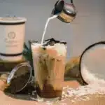 a person adding milk to the coconut coffee, with coconut placed on side