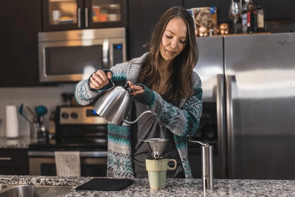 a lady making coffee holding a kettle standing in a kitchen